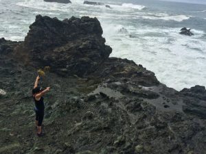 Woman raising arms up in front of white water waves on rocky shore