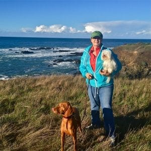 Woman in blue wind breaker holds maltese in arm and vizsla by leash on the ocean bluff trail in sea ranch