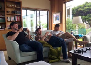 3 young men engrossed in their electronic devices while sitting together on a couch with the natural beauty of Sea Ranch showing through the windows