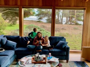 Mother, son and daughter on blue living room sofa. Boy looks out to ocean and field below, mother and daughter work on knitting. Coffee table has coloring books.