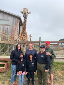 Family visits B Bryan Preserve: Mother with 7 year old boy and girl, Grandmother, Grandfather, adult son with Down syndrome, Giraffe looks over them in background