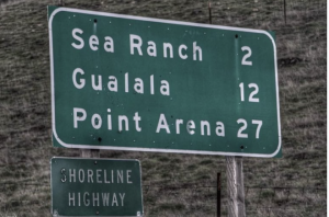 Highway road sign showing miles to Sea Ranch (2), Gualala (12), Point Arena (37) and has Shoreline Highway name under.