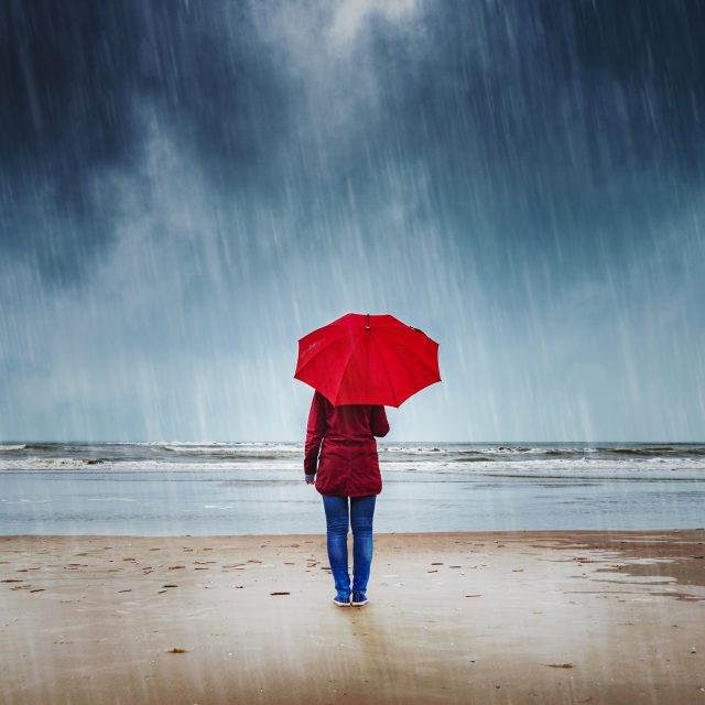 Lone woman with red umbrella is standing in the rain watching the sea
