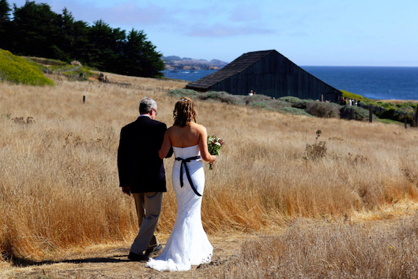 Father of the bride walking bride across the meadow trail towards the old barn at The Sea Ranch Lodge. Blue ocean in background beyond